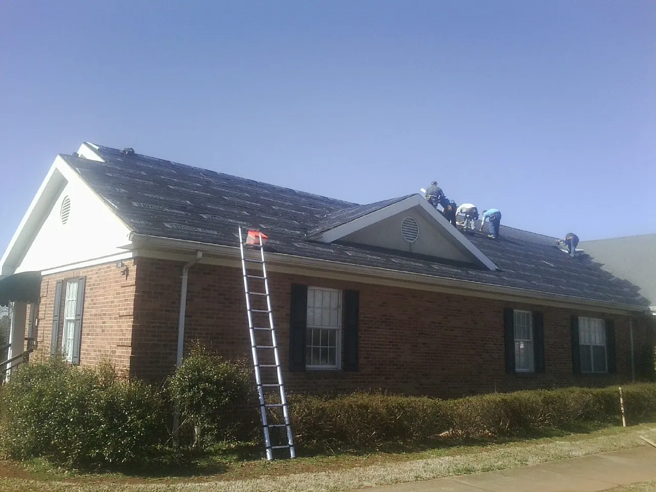 Crew tearing shingles off a brick ranch roof with a ladder set — Emergency Roof Repair work by our crew in Dickey