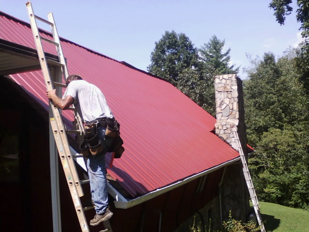 Roofer on a ladder beside a red metal roof and stone chimney — Soffit & Fascia Repair by Roofing Brothers in Dickey